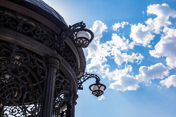 Fototapeta na wymiar Elements of a forged rotunda with lanterns against a sky with clouds. Rotunda made of black cast iron with copy space. Architectural element.