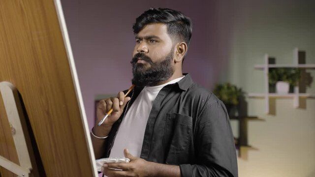 young indian beard artist thinking while painting on canvas board at home - concept of hobbies, leisure activity and professional artist.