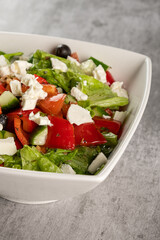 Healthy vegetable Greek salad of fresh tomatoes, red pepper, lettuce leaves, cucumbers, feta cheese and olives in a plate on a gray background. Dietary vegetarian menu. Vertical orientation, close-up