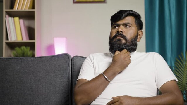 young indian beardman thinking or day dreaming while sitting on sofa at home - concept of thoughtful, wasting of time and doubtful.