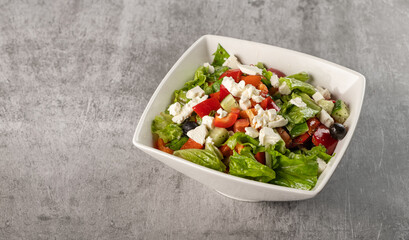 Healthy vegetable Greek salad of fresh tomatoes, red pepper, lettuce leaves, cucumbers, feta cheese and olives in a plate on a gray background. Dietary vegetarian menu. 