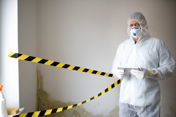 Man with white protective suit and mouth nose mask stands in front of mold on wall and works with...