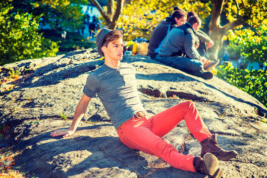 Outing In Autumn. Dressing In A Gray Long Sleeves With Roll-tab Henley Shirt, Red Jeans, Brown Boot Shoes, Wearing A Fedora Hat, A Young Handsome Man Is Sitting On Rocks Under Sunshine, Relaxing.