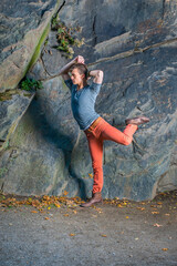 Wearing a gray Henley shirt,  red jeans and brown leather boot shoes, a young guy is stretching arms and legs, walking like a robot in the front of rocky wall..