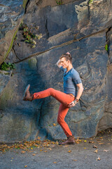 Dressing in a gray long sleeves with roll-tab Henley shirt,  red jeans, brown leather boot shoes, a young guy is stretching legs, walking robot likely by rocks..