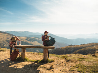 Fototapeta na wymiar Couple of hikers with backpacks enjoying valley landscape view from top of a mountain. Happy Young adult tourists, man and woman decided sit on a bench on mountain pass. Panoramic view of mountain