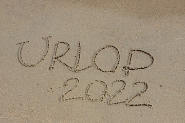 The inscription urlop 2022 in Polish, vacation 2022, on the sand by the water and the rising wave, seashore beach vacation by the sea - obrazy, fototapety, plakaty