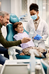 Happy boy looking his teeth in mirror while being with his father at dentist's.