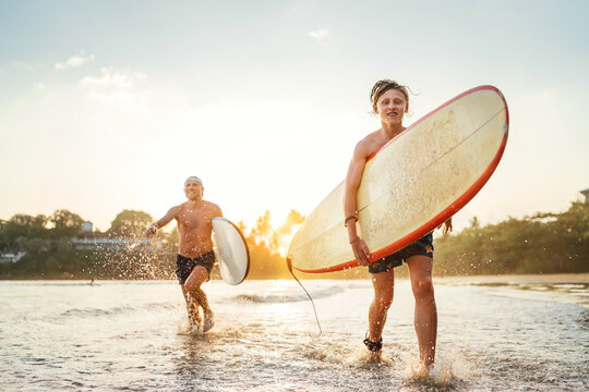 Young teen boy with a surfboard running with his father by ocean sandy beach after surfing. They are smiling and enjoying a beautiful sunset light. Family active vacation concept.