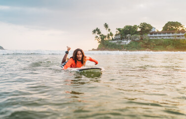 Black long-haired man paddling on long surfboard to the surfing spot in Indian ocean. Palm grove...