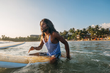 Black long-haired teen man portrait floating on long surfboard in sleeveless telnyashka, waiting...