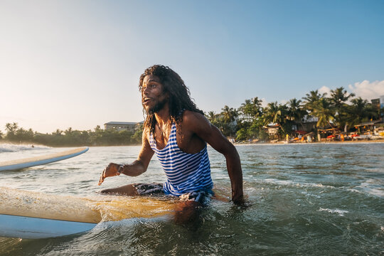 Black long-haired teen man portrait floating on long surfboard in sleeveless telnyashka, waiting for a wave ready for surfing. Extreme water sports and traveling to exotic countries concept.