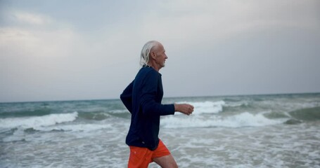 An elderly man is jogging on the beach. A gray-haired athlete runs along the beach at sunset. 4k slow motion video - Powered by Adobe
