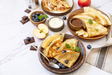 Celebrating Pancake day, healthy breakfast. Homemade sweet crepes with blueberries, ricotta, apples, bananas and chocolate on a marble tabletop. Copy space.