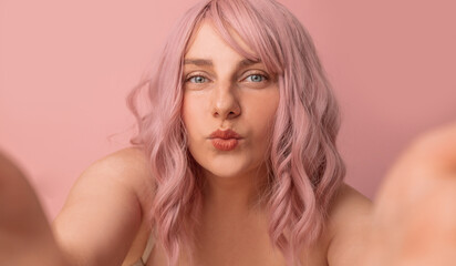 Portrait of pretty sexy girl with pink curly hair in lingerie gives an air kiss looking at camera posing isolated on pink background - obrazy, fototapety, plakaty