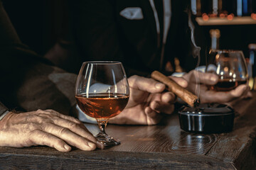 Man's hands with a cigar, elegant glass of brandy on the bar counter. Alcoholic drinks, cognac, whiskey, port, brandy, rum, scotch, bourbon. Vintage wooden table in a pub at night