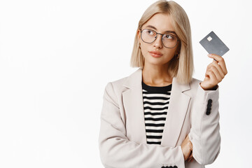 Image of business woman with thoughtful face looking away, holding credit card, deciding what to...