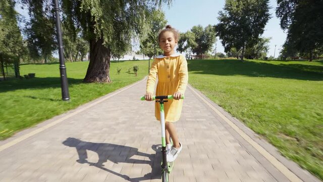 Cinematic video of a little girl traveling on her scooter in a park. American child going outdoor and having fun o a beautiful sunny day. Concept about childhood and lifestyle