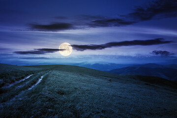 country road through alpine meadow at night. beautiful nature landscape in summer. scenery with open view in to the distant ridge and valley in full moon light. sky with clouds above the horizon