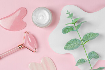 Roller jade facial massager and open jar of moisturizer on pink background with geometric shapes. Gua Sha face massage tools on pink background with green leaves