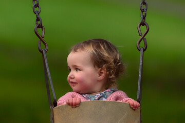 Happy curly haired blonde toddler girl in swing at park with copy space