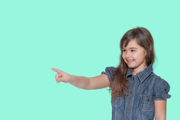 Smiling tanned long haired little girl iwears  denim costume is pointing her finger at a blank space ready for your text isolated on cyan background.