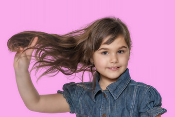 Tanned little girl wears  denim costume is showing her long hair. Isolated on pink background.