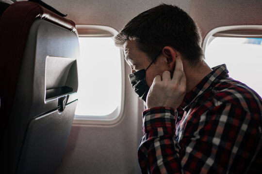 man corrects a black protective mask and plaid shirt by the window on the plane.