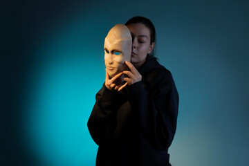Hiding behind a mask, a young woman in a dark hoodie hides her face with a mask, self-identification problems and impostor syndrome. Portrait in the studio on a dark gray background.