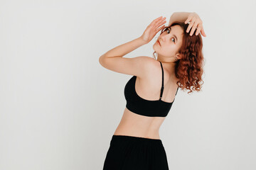 Beautiful curly redhead woman posing in black sportswear, turning around to look at the camera, on white studio background