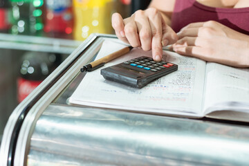 hand detail of latina woman using her small calculator and eco-friendly cardboard pen on a large notebook. doing calculations on sales and statistics in her grocery store.