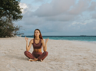 Fototapeta na wymiar A girl with long hairs in a special sports suit meditates in the lotus position on the beach of exotic tropical island. Healthy lifestyle. Travel content