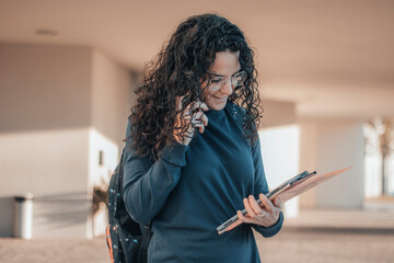 Muchacha Estudiante de cabello rizado oscuro estudiando y revisando el su teléfono móvil los apuntes del instituto en la entrada de su centro escolar antes de asistir al aula de estudios