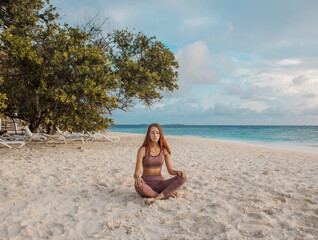 A girl with long hairs in a special sports suit meditates near the ocean on the beach of exotic tropical island. Healthy lifestyle. Mental health. Travel content