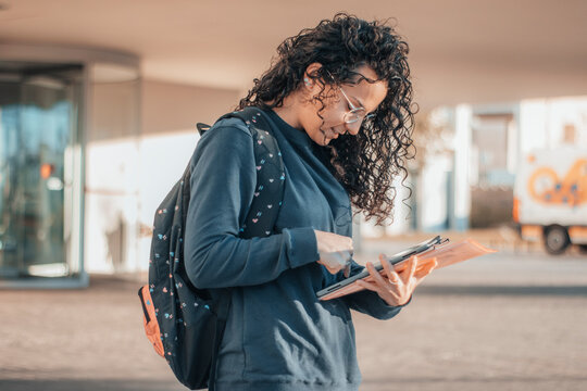 Muchacha Estudiante de cabello rizado oscuro estudiando y revisando el su tel&eacute;fono m&oacute;vil los apuntes del instituto en la entrada de su centro escolar antes de asistir al aula de estudios