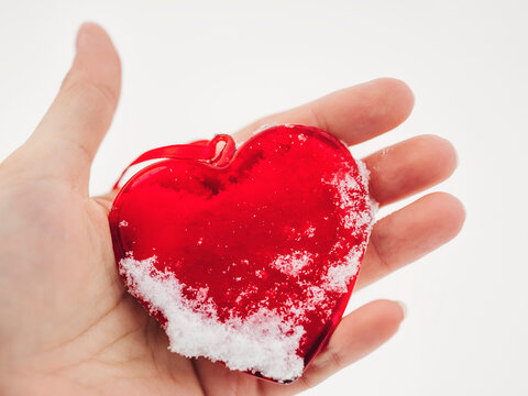 Outdoors close up image of woman hand holding a beautiful glassy red heart powdered with snow.  In a snow winter background. Love and St. Valentine day concept. Frozen heart