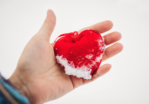 Outdoors close up image of woman hand holding a beautiful glassy red heart powdered with snow.  In a snow winter background. Love and St. Valentine day concept. Frozen heart