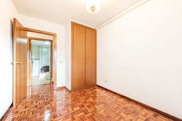 Empty room with reddish oak wood floor, built-in wardrobe with two wooden doors and toilet in the background
