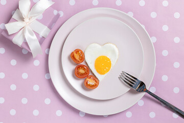 Fried egg in the shape of a heart with cherry tomatoes on a pink plate and a pink background with a gift.
