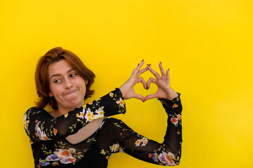 Young Latin American woman (23) looks to her right side making a doubtful gesture with her mouth, while with the fingers of her hands she makes a heart. Yellow background. Valentine's Day concepts, ge