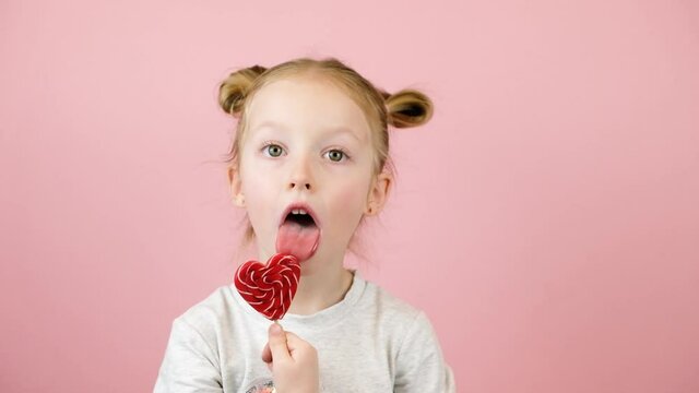 Funny little blonde girl smiling and licking red heart shape lollipop caramel on pink background. Valentines Day or Mothers Day concept - Powered by Adobe