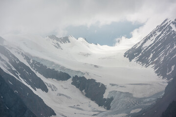 Atmospheric mountain landscape with silhouettes of sharp rocks and sunlit glacier in low clouds at changeable weather. Dramatic view to large glacier tongue in snow mountains in sunlight in low clouds