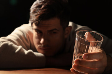 Addicted man at wooden table against black background, focus on glass of alcoholic drink