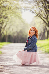 Active and happy woman in spring park. Young redhaired girl on background of green trees lane. Redhaired girl in peach skirt and jeans jacket and sneakers walks in nature. Young early spring