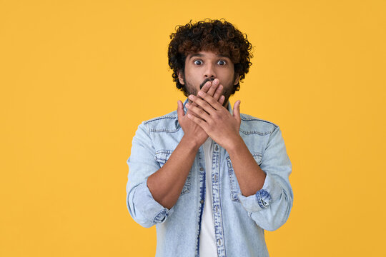 Shocked surprised young indian man looking at camera with omg face expression covering mouth with hands feeling amazed speachless silent reaction standing isolated on yellow background.