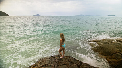 Woman chill relax resting on rock of sea reef stone, stormy cloudy ocean. Woman in swimsuit dress tunic. Concept rest tropical outdoor tourism