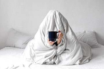 A woman wrapped in a blanket sits in bed with a cup of coffee in her hands.
