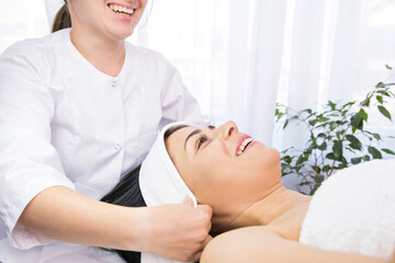 Obraz na płótnie Canvas Cosmetologist laughing putting white headband towel on patient woman head for the face rejuvenation procedures in a beauty clinic.