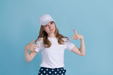 Obraz na płótnie Canvas Young caucasian girl in delivery uniform and white cap looks confident with smile on her face, pointing her fingers proudly and happily, on a blue background. Jobs for young people in advertising