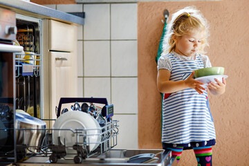 Little adorable cute toddler girl helping to unload dishwasher. Funny happy child standing in the...